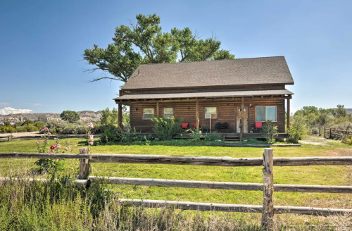 A rustic log cabin surrounded by greenery and a wooden fence under a clear blue sky.